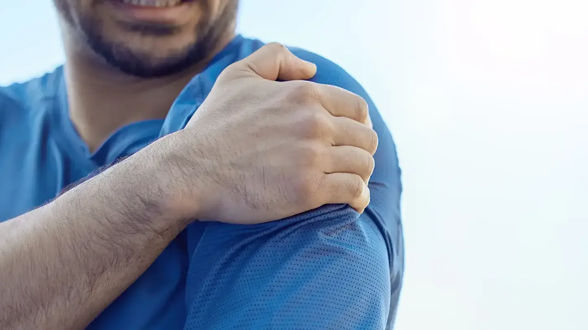 A man in a blue shirt gripping his shoulder, the kind of shoulder pain treated at HJ Manhattan Physical Therapy in New York